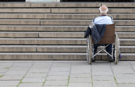 Alter Mann steht mit einem Rollstuhl vor einer Treppe und benoetigt Hilfe.